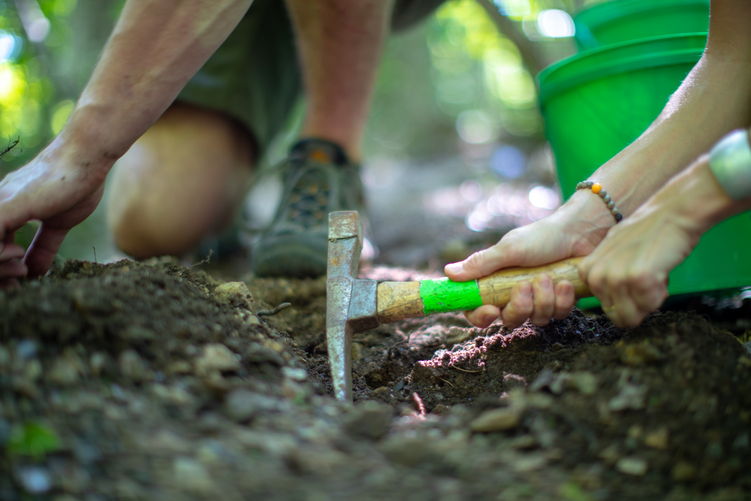 People rockhounding, a person has a pick in the dirt and another is kneeling behind them. A green bucket is in the background.