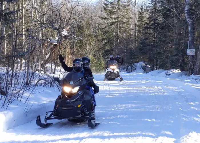 People riding on two snowmobiles on a snow covered trail in the woods