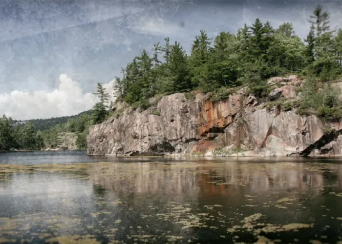 Photo of a lake, face of canadian shield in background with trees on top, sky and clouds