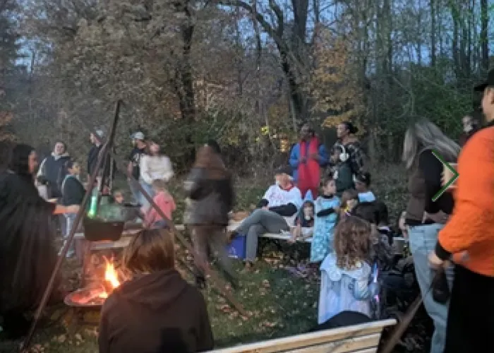 Group of people standing and sitting outside around a fire, trees in background, leaves on ground