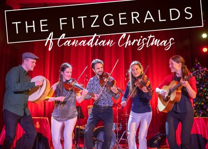 Five musicians on stage decorated with christmas decorations, three holding violins, one a drum, and one a guitar
