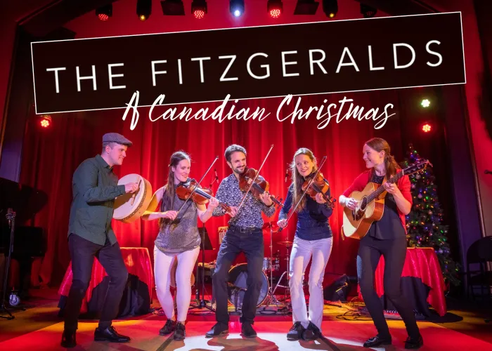 Five people on a stage decorated with christmas decor, three people are holding violines, one a drum, and the other a guitar