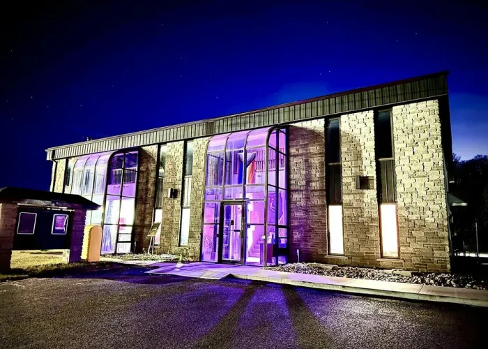 Night photo of brick building with two large entries with windows from floor to ceiling, gravel parking lot, purple/pink lighting and a pride flag can be seen through windows