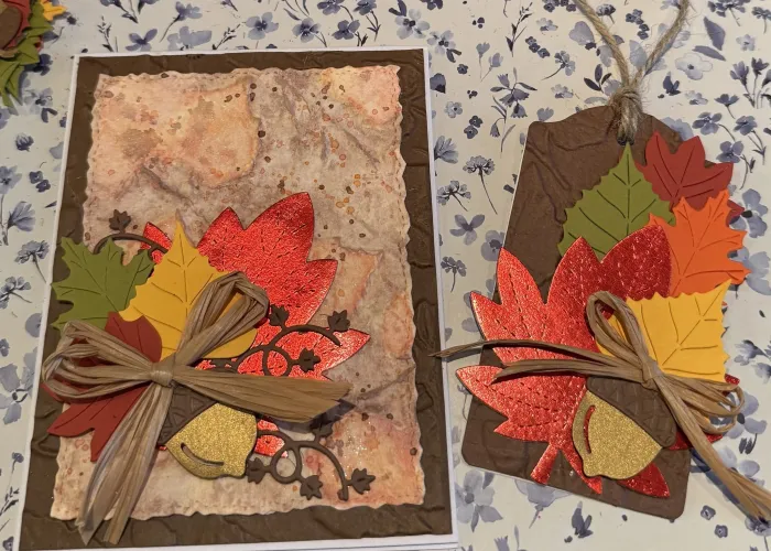 Photo of two fall-themed cards decorated with leaves, acorns, and twine on a surface patterned with blue flowers