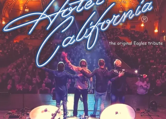 Four members of band on stage facing away towards crowd in their seats at theatre, drum set in foreground, event title written on top